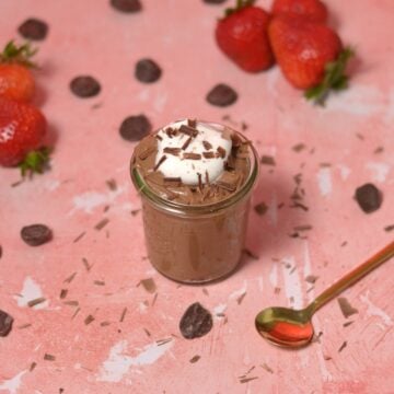 A small glass jar of chocolate mousse topped with whipped cream and chocolate shavings sits on a pink surface, surrounded by strawberries, chocolate pieces, and a gold spoon.