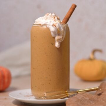 A tall glass filled with a creamy pumpkin smoothie, topped with whipped cream, sprinkled cinnamon, and a cinnamon stick. Small pumpkins and a mini whisk are nearby on a plate.