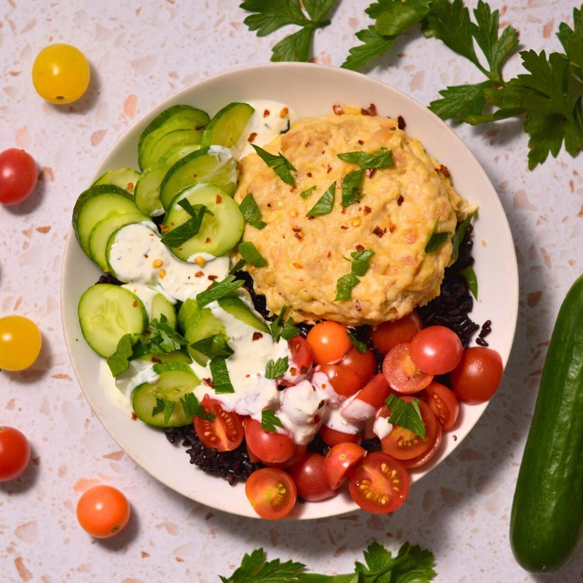A bowl with black rice, sliced cucumbers, cherry tomatoes, creamy hummus, and herb yogurt sauce, garnished with chopped parsley. Extra tomatoes, cucumber, and parsley surround the bowl on a light surface.