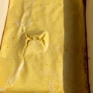 A close-up of a baked dish with a golden-brown surface, featuring a noticeable crack and uneven texture in the center—reminiscent of a classic Dutch baby. The lighting highlights its smooth and airy top.