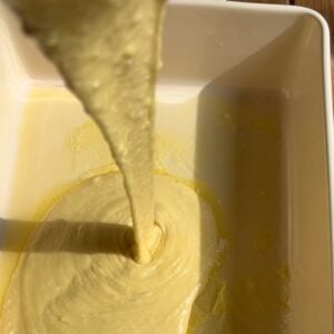 Thick yellow dutch baby batter being poured into a white rectangular baking dish, with sunlight casting soft shadows on the surface.