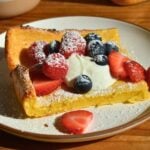 A baked Dutch baby pancake topped with whipped cream, sliced strawberries, blueberries, raspberries, and a dusting of powdered sugar on a white plate.