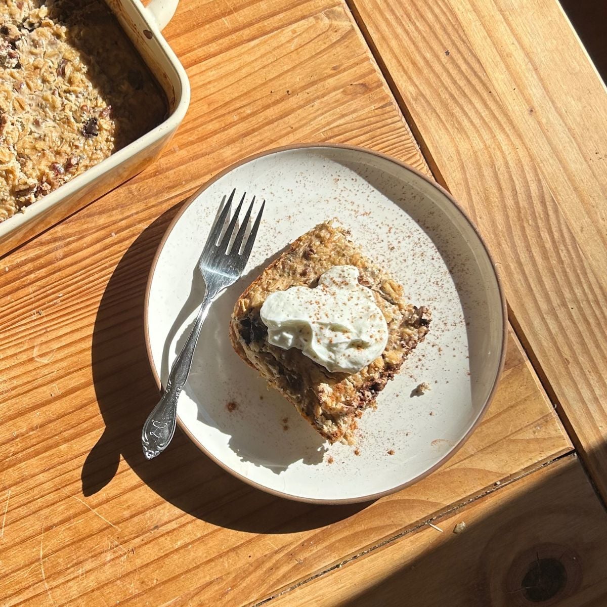 A slice of banana bread baked oats topped with a dollop of whipped cream sits on a white plate with a fork. Sunlight shines on the wooden table beneath, and a baking dish with more baked oats is partially visible.