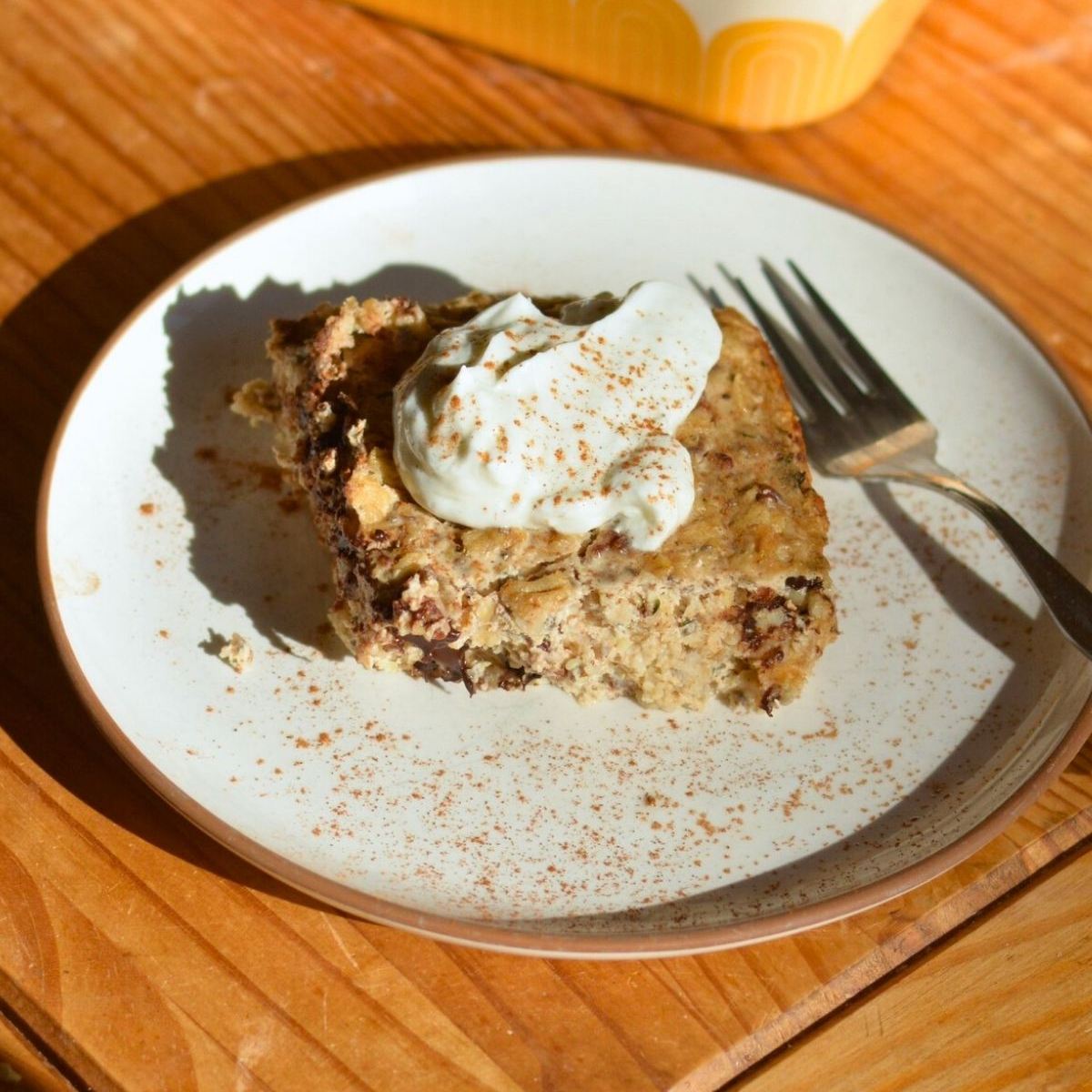 A slice of baked oatmeal with chocolate chips, topped with whipped cream and a sprinkle of cinnamon, sits on a white plate with a fork on a wooden table.