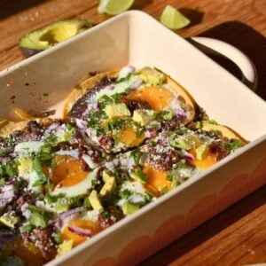 A casserole dish with huevos rancheros consisting of egg, refried beans, avocado, onion, and cilantro.