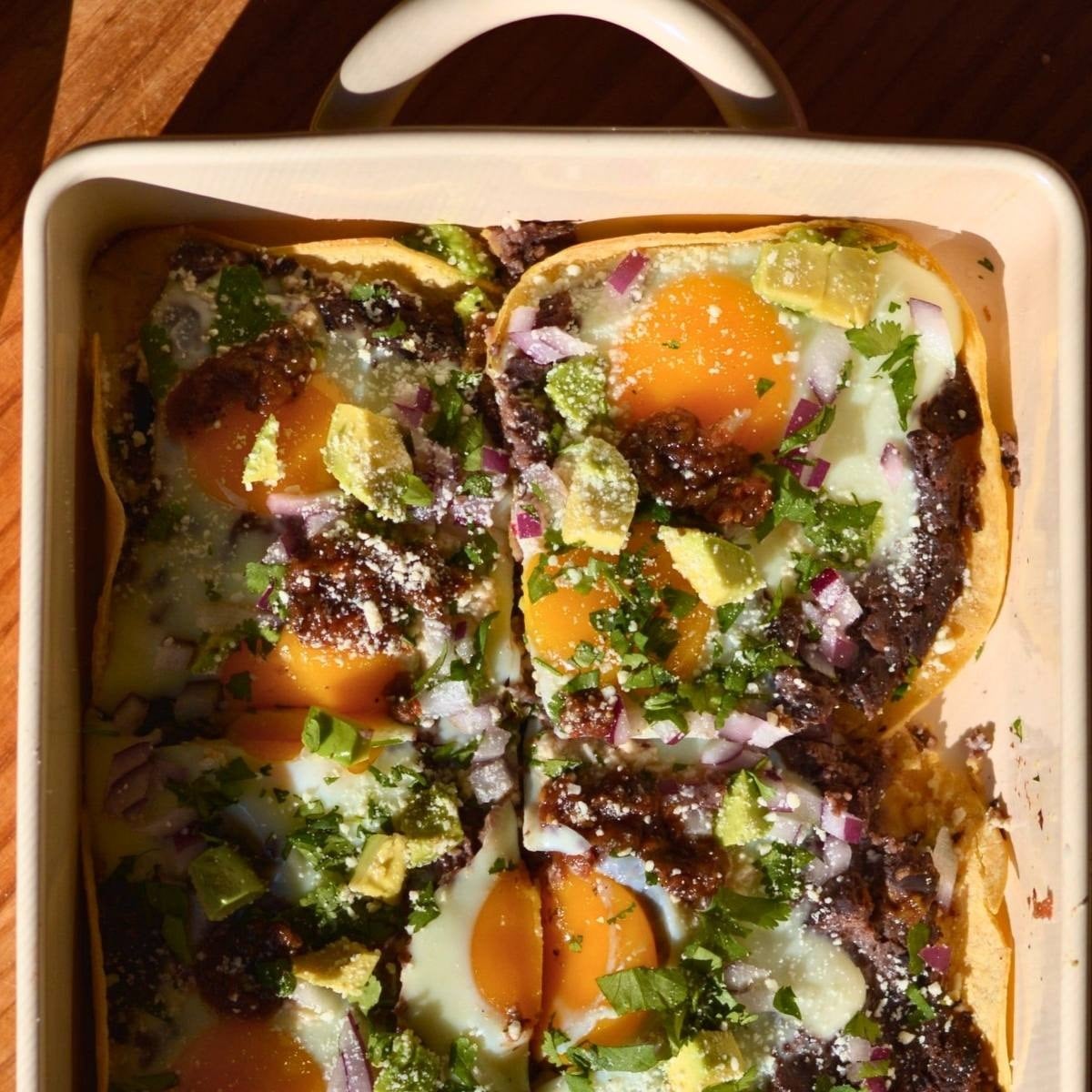 A casserole dish with huevos rancheros consisting of egg, refried beans, avocado, onion, and cilantro.