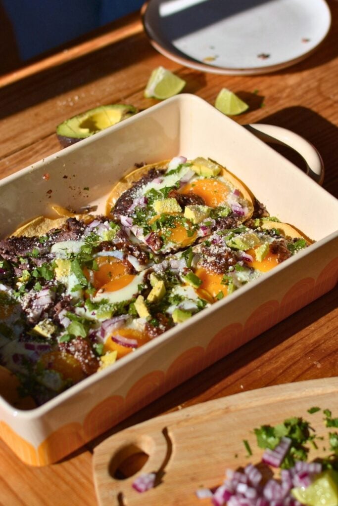 A casserole dish with huevos rancheros consisting of egg, refried beans, avocado, onion, and cilantro.