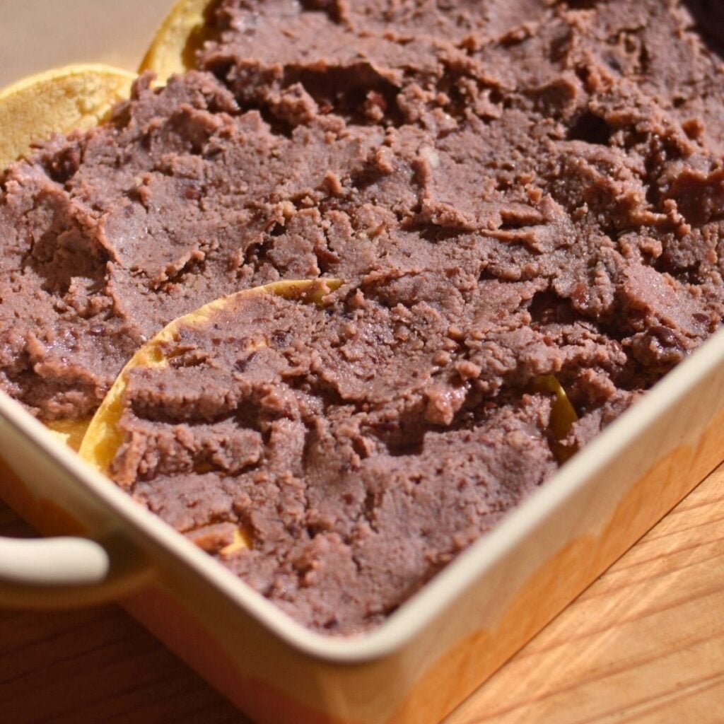A casserole dish with refried black beans on top of corn tortillas.