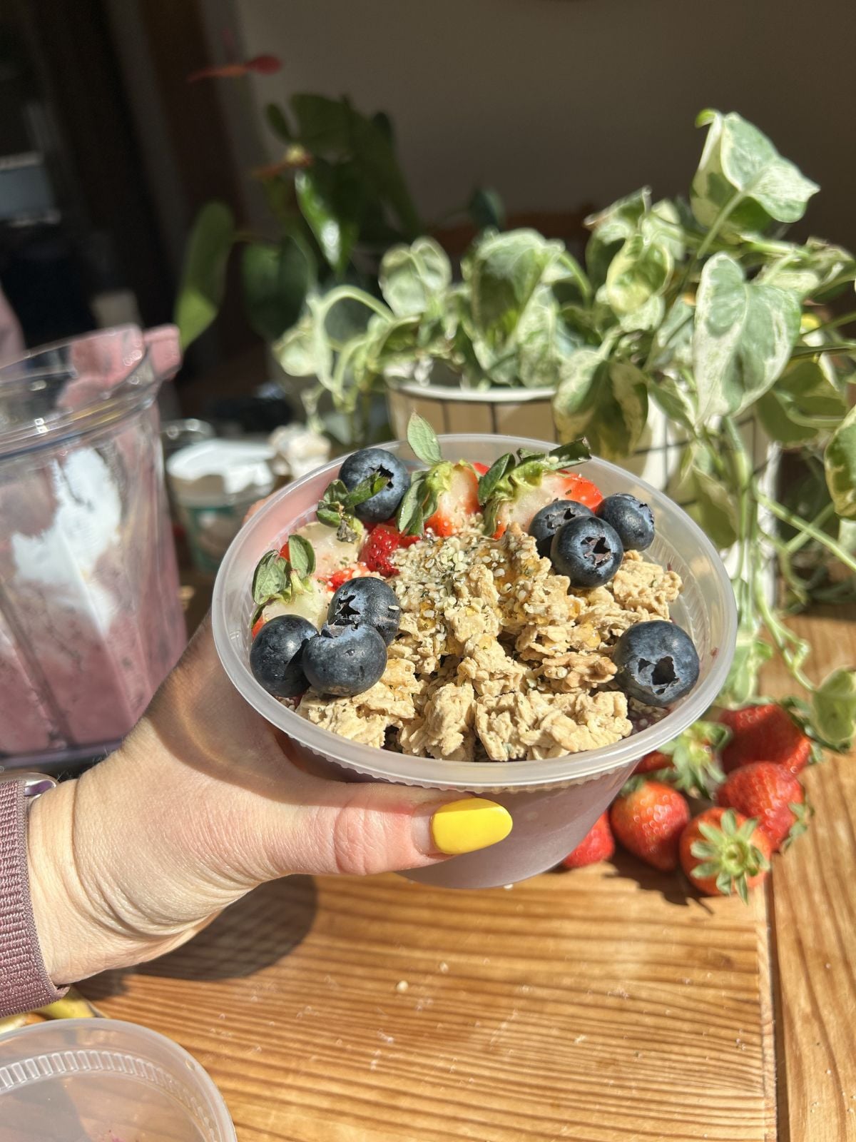 A hand holding a protein acai bowl in a plastic tub with granola and berries on top.