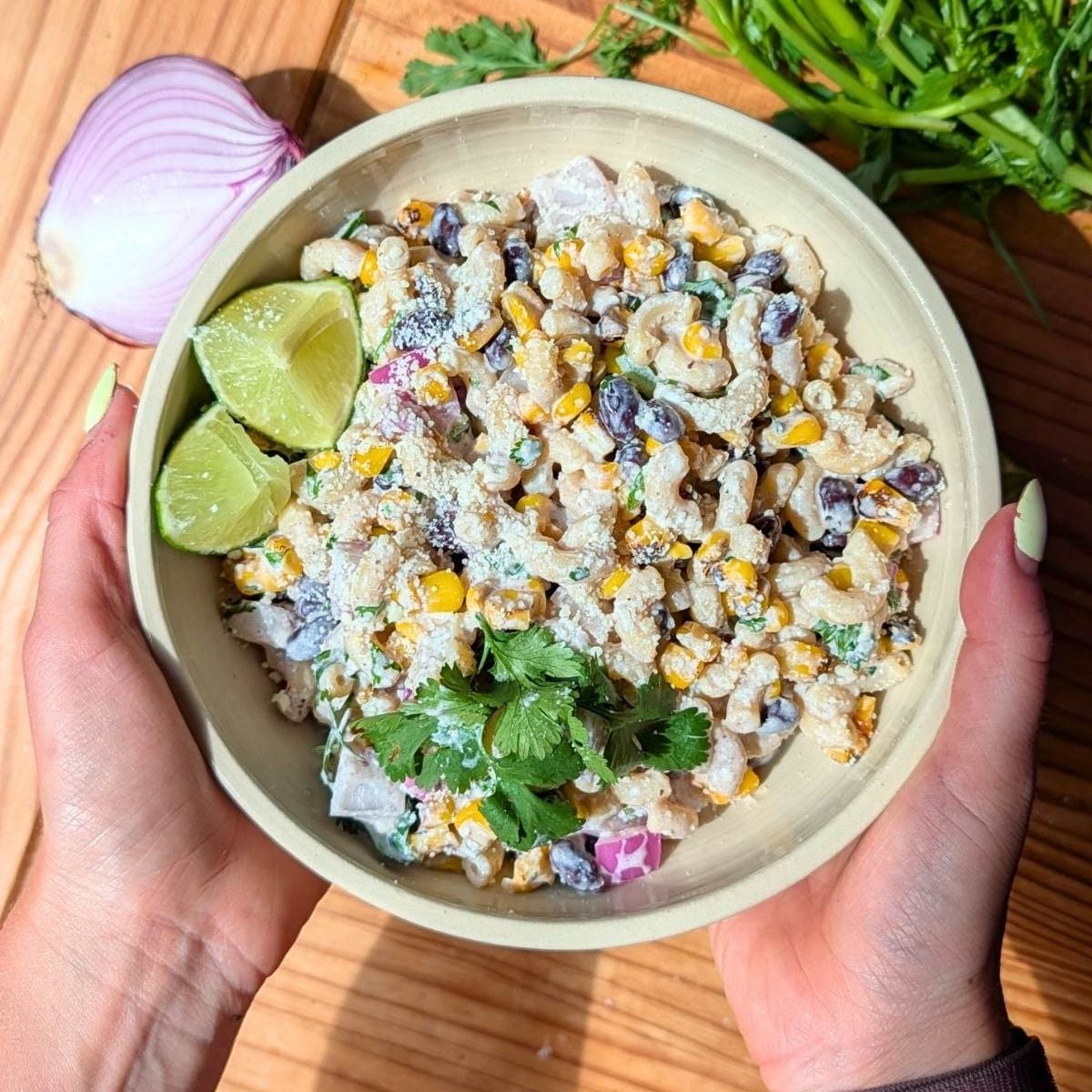 Hands holding a cream colored bowl filled with street corn pasta salad with cilantro and lime wedges.