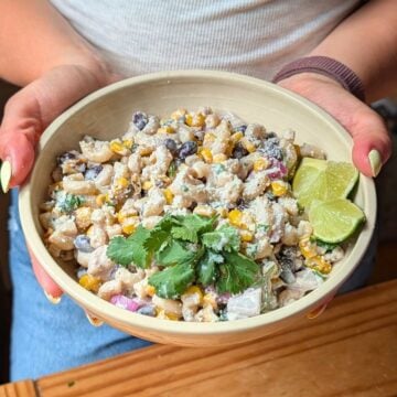 Hands holding a cream colored bowl filled with street corn pasta salad with cilantro and lime wedges.