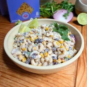A cream colored bowl filled with street corn pasta salad with cilantro and lime wedges.