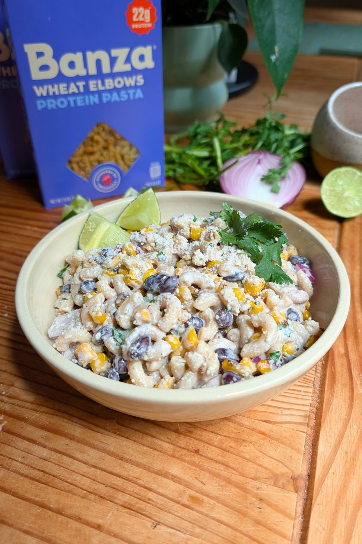 A cream colored bowl filled with street corn pasta salad with cilantro and lime wedges.